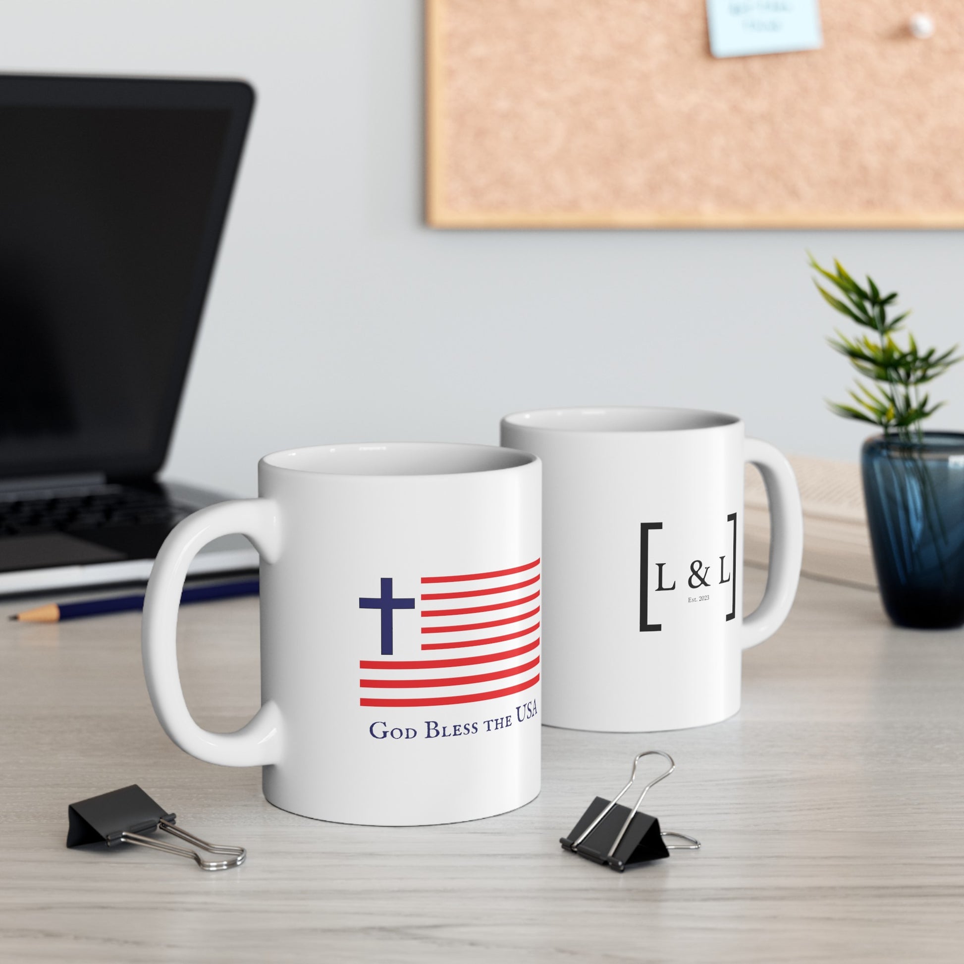 a white mug with a blue cross and an American flag with the words God Bless the USA and another mug with the for lord and liberty logo that is sitting on a desk