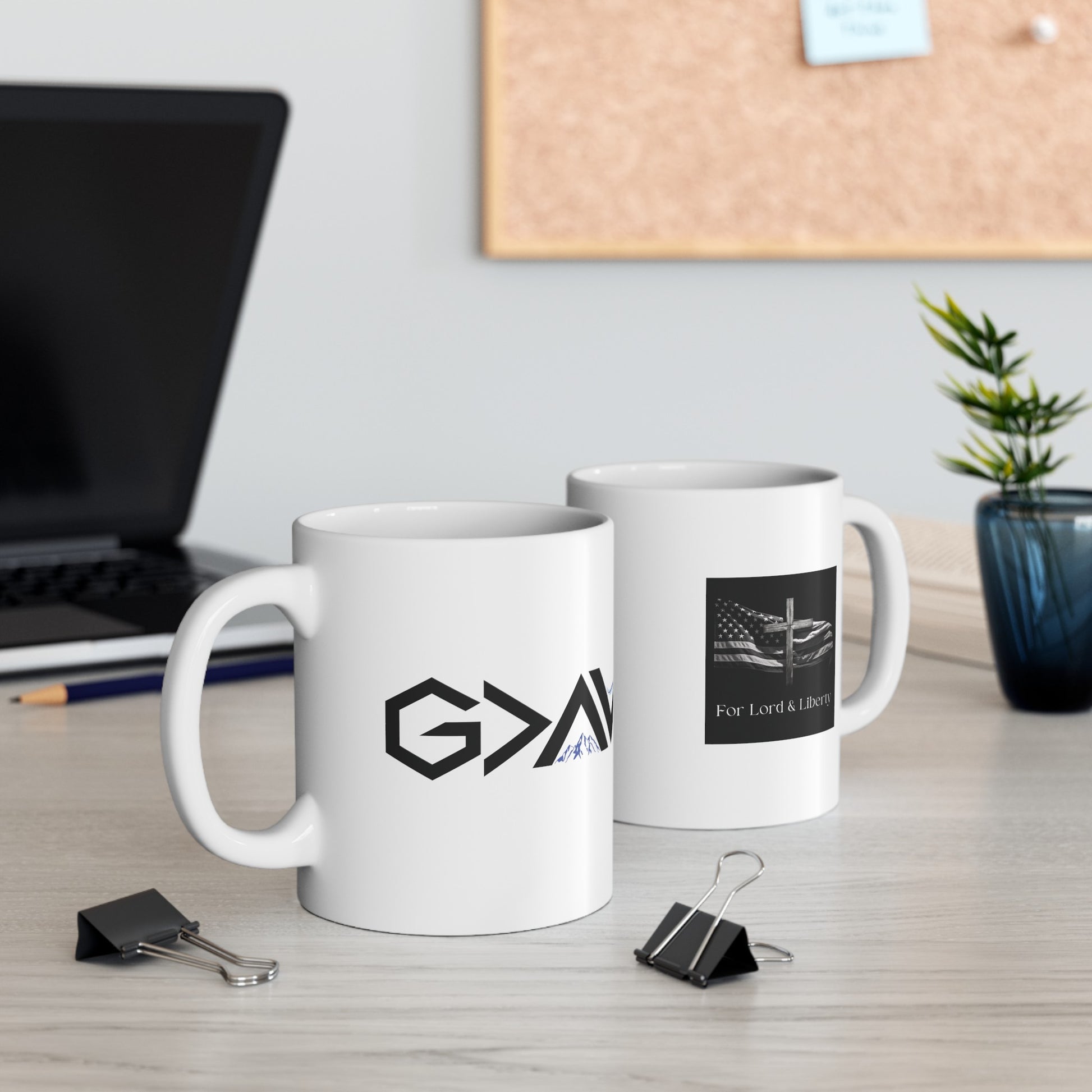 a white mug that say God is Greater than the Mountains and Valleys and another mug with the for lord and liberty logo that is sitting on a desk