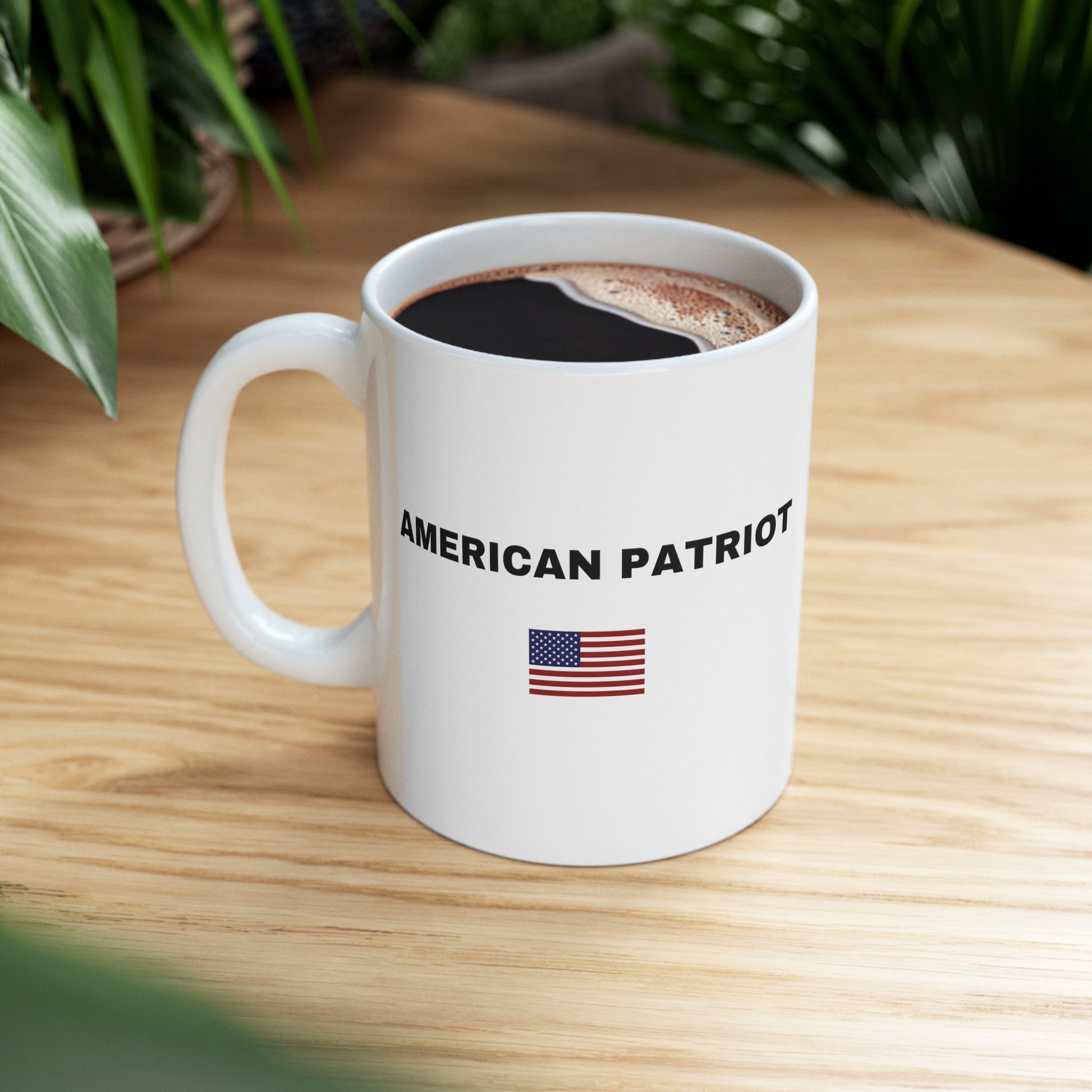 a white mug with the words American Patriot in black with an American Flag on the front sitting on a desk