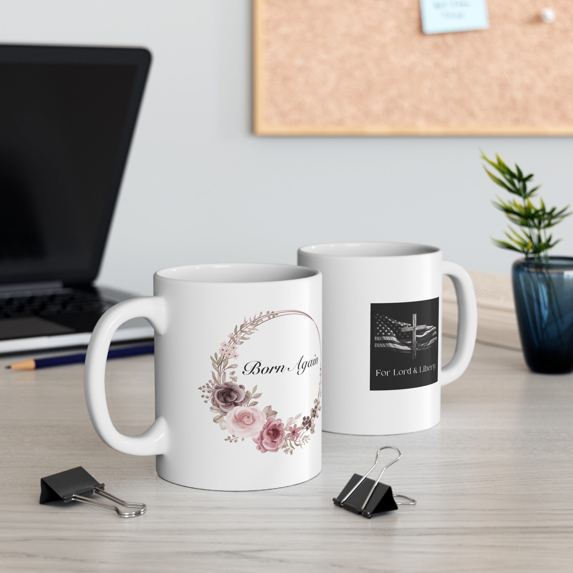 a white mug with the words Born Again with a flower wreath and another white mug with the for lord and liberty logo that is sitting on a desk