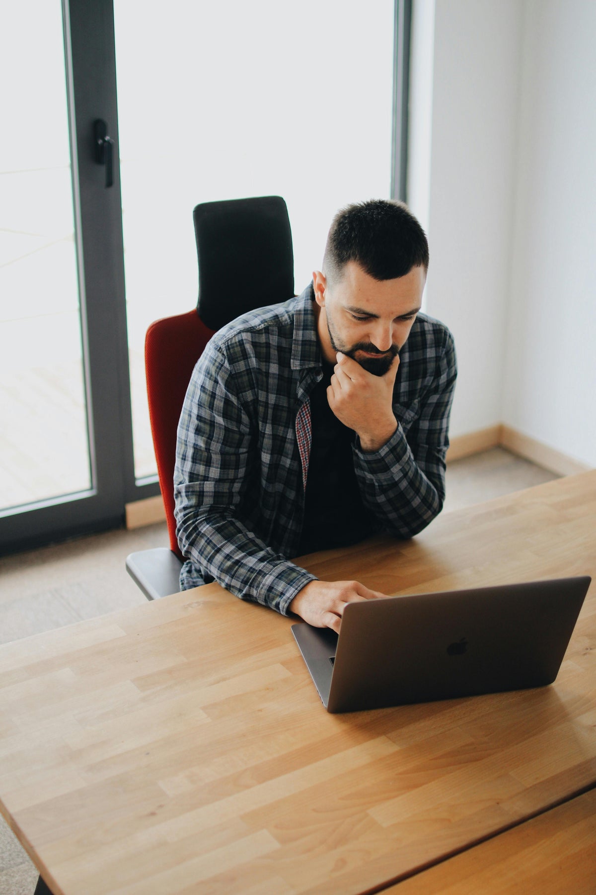 Man in a flannel shirt deep in thought while working on a laptop at a wooden table
